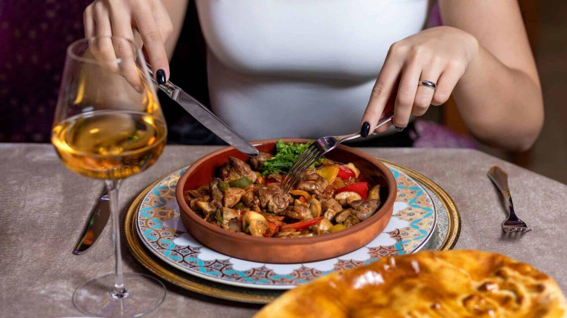 A lady eating a bowl of food she has a white top on the food is in a brown bowl on a blue and white Greek style plate