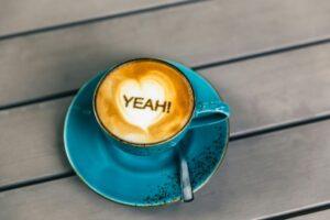 A blue cappuccino cup sits on a blue wooden planed counter top with latte art on top, the art features a "Heart" and the word "Yeah" signifying its important to celebrate the smallest of wind in you battle against FIbromyalgia and chronice pain.